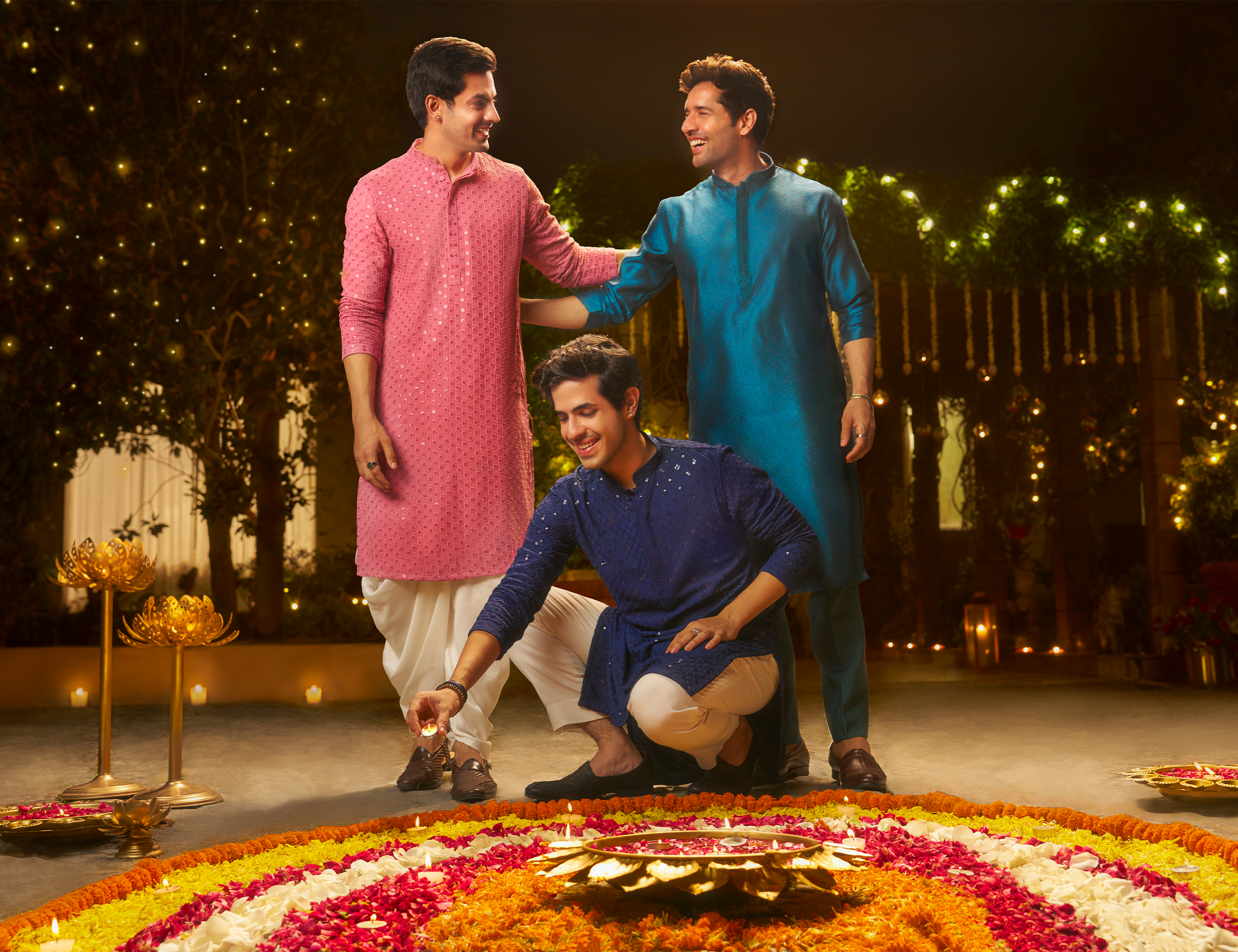 Men in traditional blue, purple and pink kurta applying diya in flower rangoli